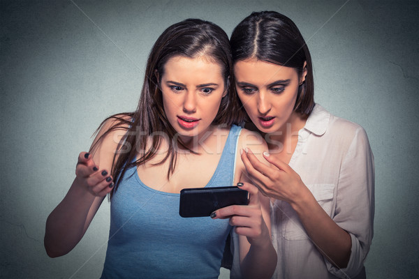 Stock photo: Two surprised girls looking at cell phone discussing latest gossip news