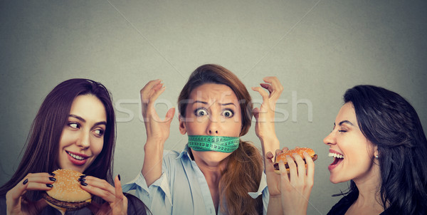 Two happy women eating hamburgers looking at stressed girl with measuring tape around her mouth.  Stock photo © ichiosea