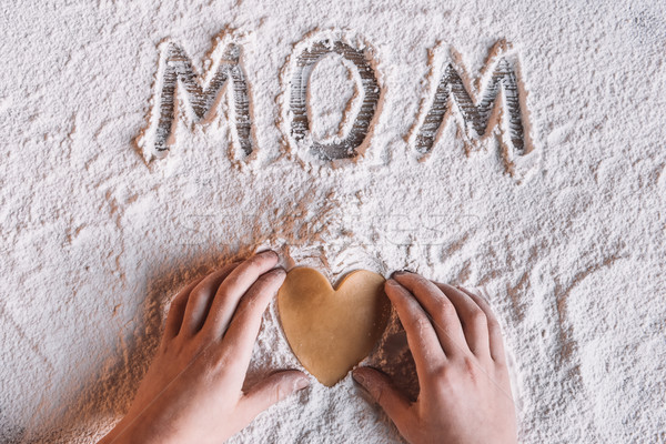 Stock photo: 'Partial top view of child holding heart shaped cookie and word mom written in flour, Mothers day co