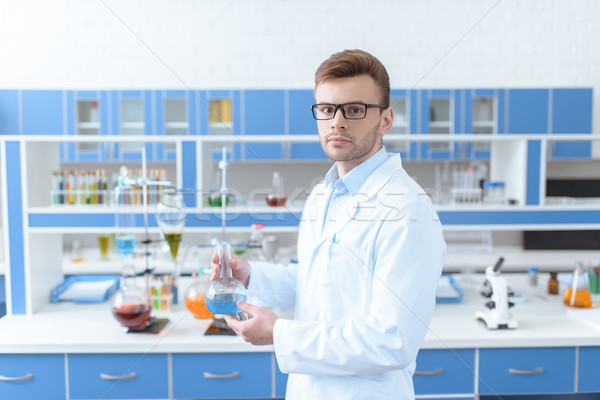 Young man scientist in eyeglasses holding flask with reagent and looking at camera Stock photo © LightFieldStudios