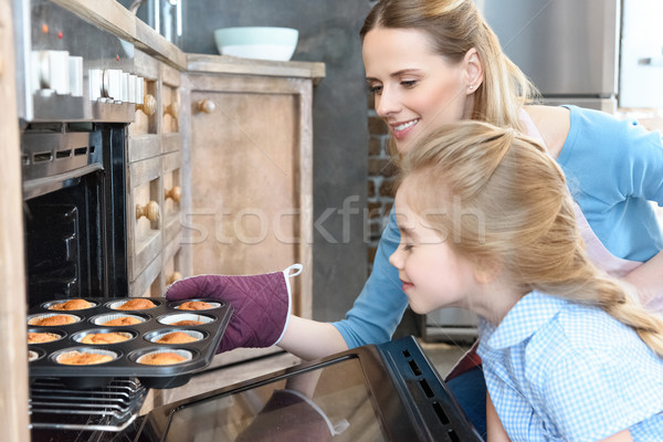Stock photo: side view of happy mother getting cupcakes from oven with daughter near by