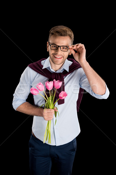 Smiling handsome man holding pink tulips and adjusting eyeglasses on black, international womens day Stock photo © LightFieldStudios