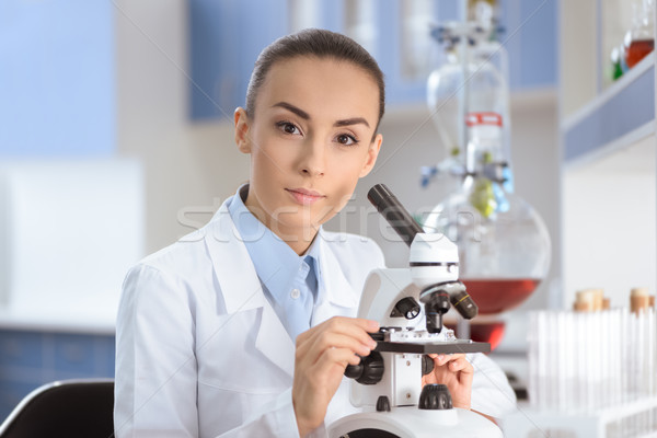 Young woman scientist in lab coat working with microscope and looking at camera Stock photo © LightFieldStudios