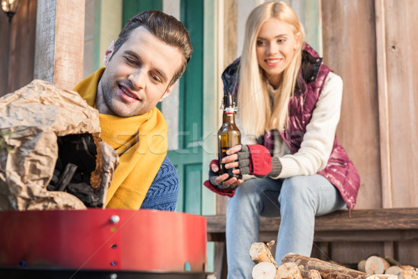 Smiling woman with beer bottle looking at handsome man filling grill with charcoal   Stock photo © LightFieldStudios