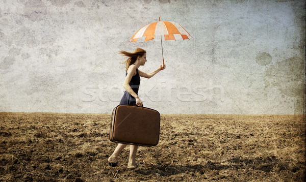 Stock photo: Redhead girl with umbrella and suitcase at windy grass meadow.