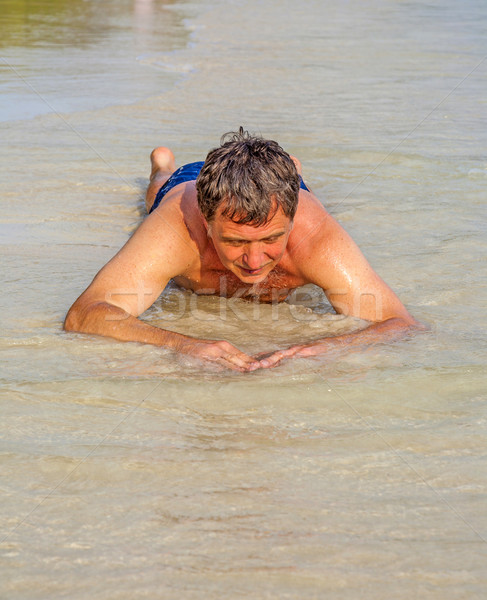 Man in bathingsuit is lying at the beach and enjoying the saltwater with tiny waves and smiles Stock photo © meinzahn