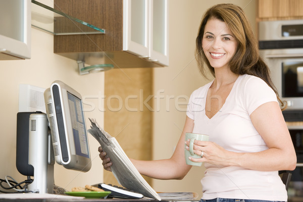 Stock photo: Woman in kitchen at computer with newspaper and coffee smiling