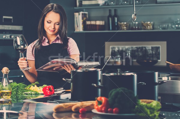 Cheerful young woman in apron on modern kitchen with cookbook and glass of wine  Stock photo © Nejron
