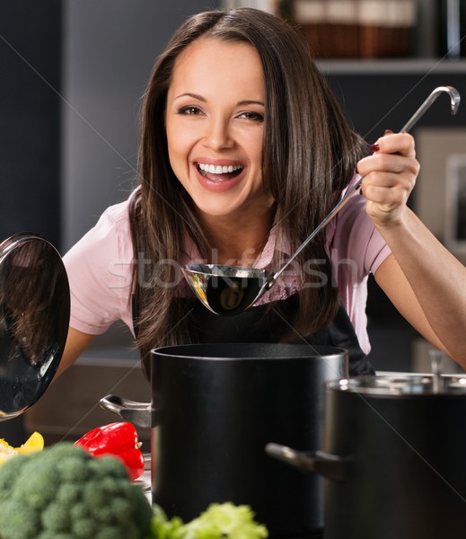 Cheerful young woman in apron on modern kitchen will ladle tasting from pot Stock photo © Nejron