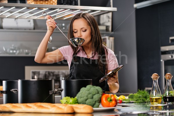 Cheerful young woman in apron on modern kitchen with ladle tasting from pot Stock photo © Nejron