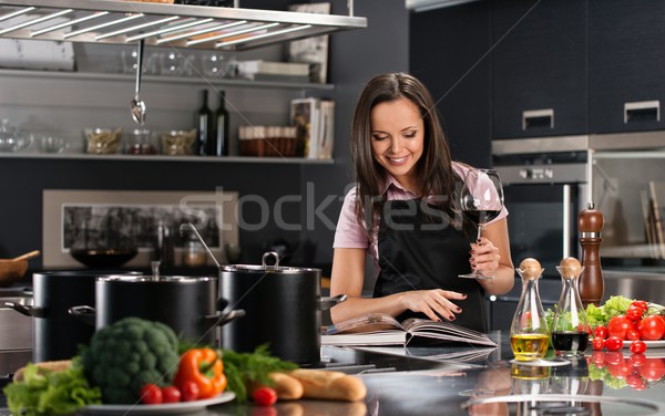Cheerful young woman in apron on modern kitchen with cookbook and glass of wine  Stock photo © Nejron