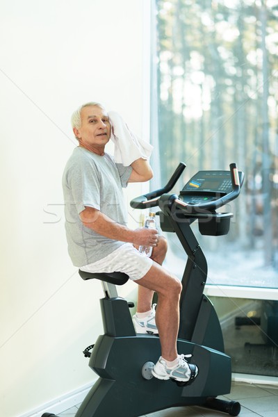 Tired senior man with towel on exercise bike in fitness club Stock photo © Nejron