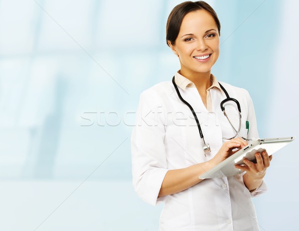 Stock photo: Young positive brunette doctor woman taking notes on tablet pc 