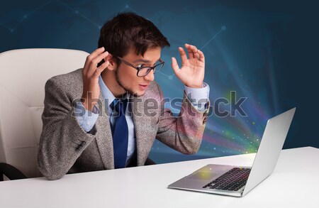 Angry young man sitting at dest and typing on laptop with message icons comming out Stock photo © ra2studio