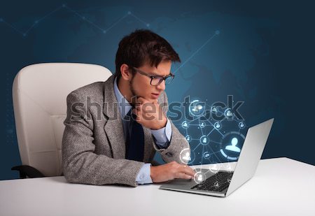 Attractive young man sitting at desk and typing on laptop with social network icons comming out Stock photo © ra2studio