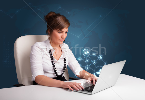Beautiful young lady sitting at desk and typing on laptop with social network icons comming out Stock photo © ra2studio