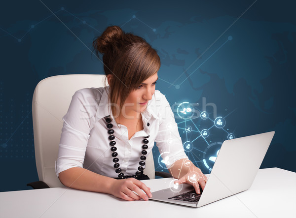 Beautiful young lady sitting at desk and typing on laptop with social network icons comming out Stock photo © ra2studio