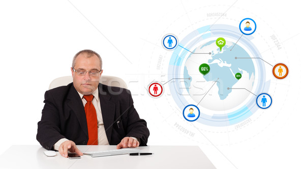 Stock photo: businessman sitting at desk and holding a mobilephone with globe and social icons, isolated on white