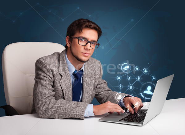 Attractive young man sitting at desk and typing on laptop with social network icons comming out Stock photo © ra2studio