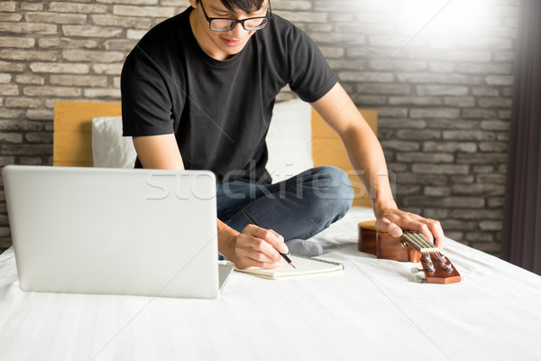 Happy young asian man playing ukulele sitting on bed in bedroom. Stock photo © snowing