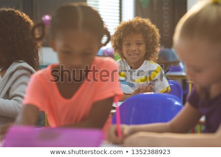 Zdjęcia stock: Front View Of Schoolboy Looking At Camera While Sitting At Desk In School With School Kids Drawing I