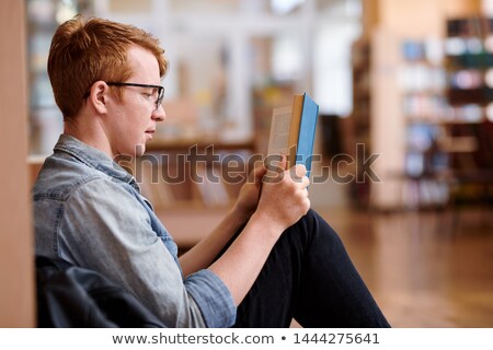 Stok fotoğraf: Young Reader In Casualwear Reading By Bookshelf In Library