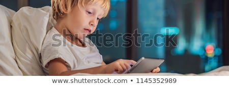 Foto d'archivio: The Boy Uses The Tablet In His Bed Before Going To Sleep On A Background Of A Night City Children A