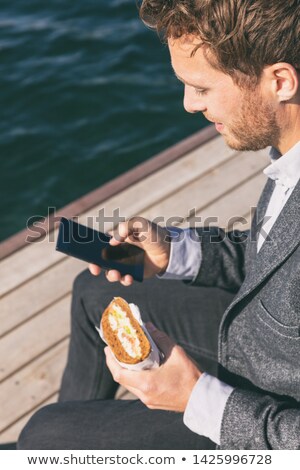 Stockfoto: Professional Man On Lunch Break From Work Using Cellphone Eating Sandwich Outside By The Canal Water
