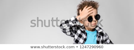 Foto stock: A Handsome Boy In A Plaid Shirt Gray Shirt And Jeans Stands On A Gray Background A Boy In Red Head