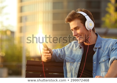 Stockfoto: Young Guy Sitting In Park Outdoors Using Mobile Phone Listening Music With Earphones