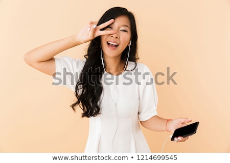 [[stock_photo]]: Happy Asian Woman Posing Isolated Over Beige Wall Background Listening Music With Earphones By Phone