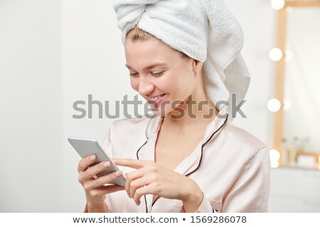 Stockfoto: Young Smiling Woman In Silk Pajamas With Towel On Head Scrolling In Smartphone