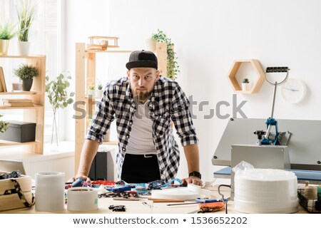 Stock photo: Young Casual Craftsman Looking At You While Bending Over Table During Work