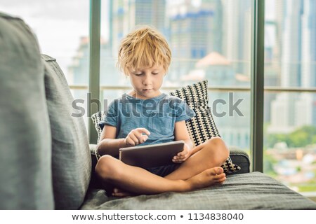 Stok fotoğraf: Boy Uses A Tablet At Home On The Couch In The Background Of A Window With Skyscrapers Modern Childr