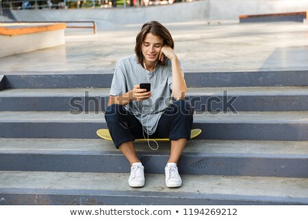 Stock fotó: Happy Oung Skater Guy Sit In The Park Listening Music With Earphones Using Phone
