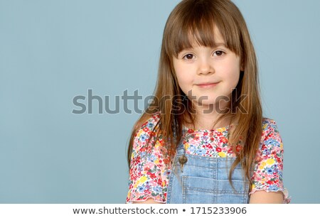 Foto d'archivio: Sweet Little Girl 6 7 Years Old Posing In Dungarees Jeans And Flower Pattern Blouse On Blue