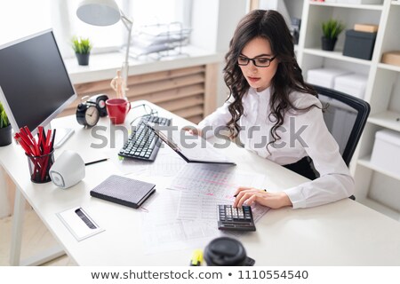 Stockfoto: A Young Girl Is Sitting At The Office Desk And Is Blessing On The Calculator