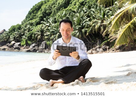 Stok fotoğraf: Young Man Sitting On The Sandy Beach Using Digital Tablet