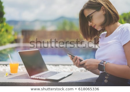 Stock photo: Young Female Freelancer Sitting Near The Pool With Her Laptop In The Hotel Browsing In Her Smartphon