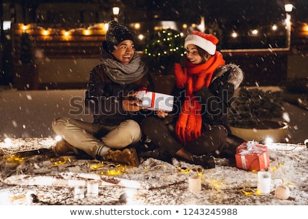 Stockfoto: Emotional Excited Young Loving Couple Sitting Outdoors In Evening In Christmas Hat Holding Gift Box