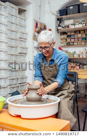 Stockfoto: Senior Female Potter Working On Pottery Wheel While Sitting In