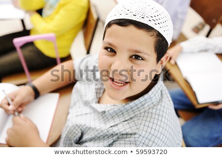 Foto stock: Very Positive Kid With White Small Hat Sitting On Desk In Classroom And Smiling