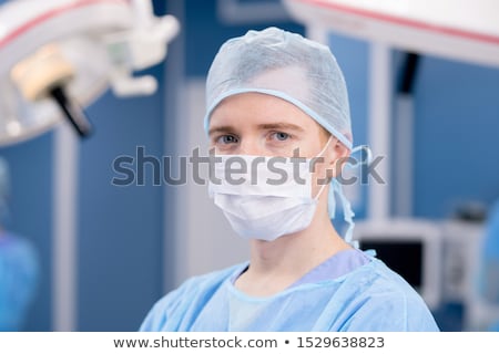 Foto stock: Young Intern Or Surgeon In Protective Mask And Uniform Looking At You