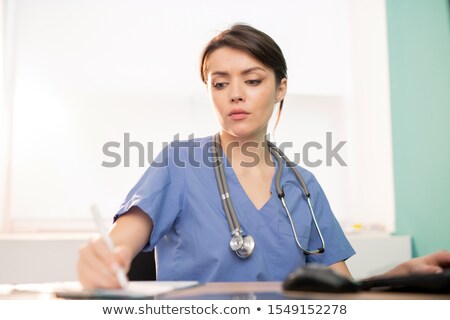 Foto stock: Young Serious Doctor Or Intern In Blue Uniform Making Working Notes By Workplace