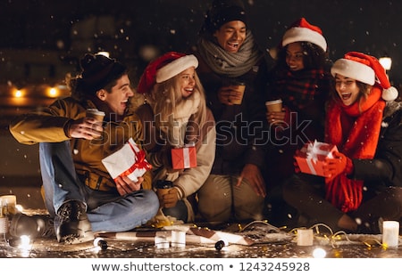 Stockfoto: Happy Young Friends Sitting Outdoors In Evening In Christmas Hats Holding Gift Box