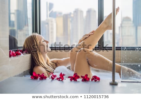 Foto stock: Young Woman Sits In A Bath With Foam And Frangipani Flowers Against The Background Of A Panoramic Wi