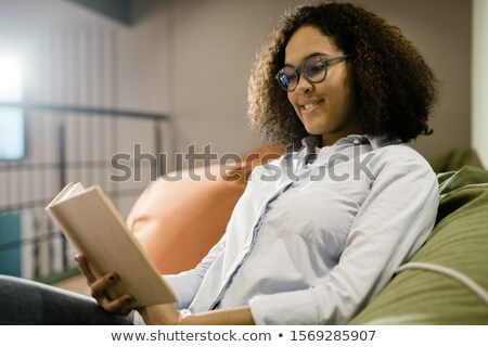 Foto stock: Happy Young Clever Female Student In New Eyeglasses Reading Book On Couch