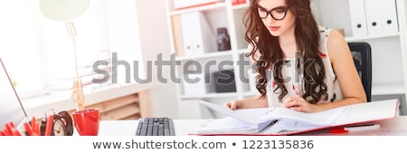 Stock photo: A Young Girl In Glasses Sits At A Table In The Office Holds A Pencil In Her Hand And Works With Doc