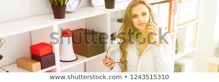 [[stock_photo]]: A Beautiful Young Girl Stands Near A Rack In The Office Talking On The Phone And Holding Glasses In