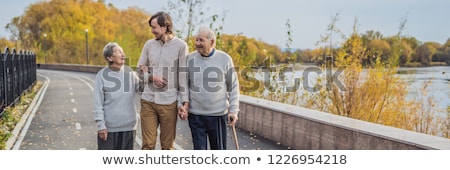Zdjęcia stock: An Elderly Couple Walks In The Park With A Male Assistant Or Adult Grandson Caring For The Elderly